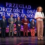 Kobieta stojąca na scenie, prezentująca się przed publicznością, z pięcioma mężczyznami w garniturach stojącymi za nią.