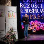 Mężczyzna w okularach stojący na scenie podczas prezentacji trzyma teczkę w ręku i mówi do mikrofonu. Obok niego znajdują się dwa długie banery prezentujące Stowarzyszenie To My oraz Środowiskowy Dom Samopomocy w Kraśniku (typ C, D).