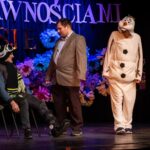 Na scenie trzech mężczyzn: jeden siedzi na krześle w zimowym stroju, drugi stoi naprzeciwko niego w garniturze, a trzeci obserwuje ich w kostiumie bałwana.