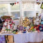 Stół wystawowy prezentujący różnorodne ręcznie wykonane przedmioty, w tym dziergane czapki, kolorowe ozdoby oraz dekoracje świąteczne. Obok stołu znajdują się obrazy. Za eksponowanymi przedmiotami umieszczona jest ramka z tekstem: 'Środowiskowy Dom Samopomocy w Puławach