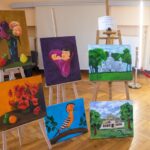 Wystawa pięciu obrazów na sztalugach: wazon z kwiatami, dziecko w kwiatku fioletowym, biały pałac z drzewami w tle, czerwone maki, ptak na gałęzi, domek wiejski.