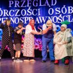 Grupa pięciu osób w kolorowych kostiumach na scenie. Kobieta w czerwonej chuście na głowie ściska dłoń mężczyzny w niebieskim kombinezonie, który ociera czoło. Przed nim siedzi kobieta w kremowej todze i czapce z zielonym pomponem. Za plecami kobiety w czerwonej chuście stoi mężczyzna w sportowym stroju i czapce z daszkiem, a za nim kolejny mężczyzna w czarnym kombinezonie w białe kropki z kapturem i maską na twarzy.