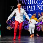 Mężczyzna przebrany za ptaka, z długim pomarańczowym dziobem i czerwonymi spodniami, tańczy na scenie. W tle występują inni artyści w różnych kostiumach zwierzęcych. Po lewej stronie częściowo widoczne jest pianino.