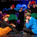 Kobieta w pomarańczowym tutu siedzi na podłodze, podczas gdy mężczyzna, ubrany w niebieską kurtkę i oliwkową czapkę, siedzi w pobliżu. W tle stoją trzy osoby w zielonym tutu, które noszą opaski ozdobne z czerwoną różą na głowie i unoszą ręce do góry.