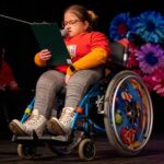 Dziewczyna w okularach, siedząca na wózku inwalidzkim, śpiewa do mikrofonu, trzymając tekst piosenki w ręku.