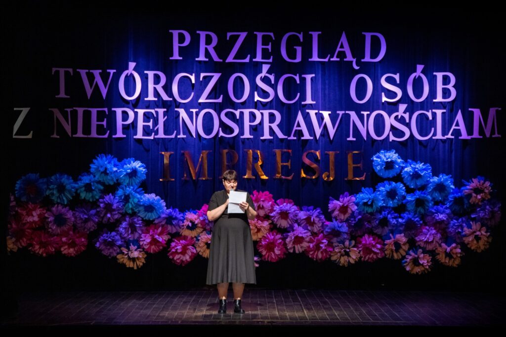 Kobieta na scenie trzyma mikrofon i kartkę. Za nią znajduje się tło z dużymi, kolorowymi kwiatami i tekstem:'Przegląd Twórczości Osób z Niepełnosprawnościami IMPRESJE'
