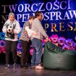 Na scenie, przed kolorowym tłem, stoją trzy osoby. Jeden z mężczyzn trzyma puf worek, jakby próbując go ciągnąć. Za nim stoi kobieta, obejmująca go i próbująca pomóc. Obok nich, kolejny mężczyzna z czapką odwróconą daszkiem do tyłu trzyma w ręku piłkę nożną.