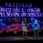 Dwie kobiety stoją na scenie i śpiewają do mikrofonów, trzymając w rękach tekst piosenki.