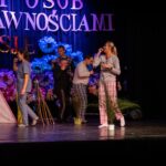 Sześciu ludzi w codziennych ubraniach porusza się po scenie, wykonując różne czynności związane z jedzeniem. Obok nich znajduje się różowy namiot tipi.