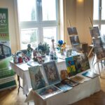 Stół wystawowy z różnymi rękodziełami i obrazami. Za stołem stojący baner z napisem: 'Dom Pomocy Społecznej w Popkowicach' z danymi kontaktowymi.