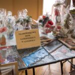 Stół wystawowy na którym są różne obrazy i rękodzieła,zapakowane w przezroczysty ceflon. Na szyldzie napis: „Środowiskowy Dom Samopomocy w Annopolu”. Na podłodze leżą dodatkowe obrazy.