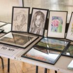 Oprawione dzieła sztuki wystawione na stole, w tym szkice ołówkiem kobiet i akwarele kwiatów. Na kartce widnieje napis „STOWARZYSZENIE TO MY”.