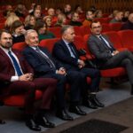 Czterech mężczyzn w garniturach siedzi na czerwonych fotelach teatralnych, skupionych na przedstawieniu. Jeden z nich patrzy w obiektyw aparatu. W tle widać innych ludzi siedzących na fotelach.