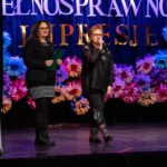 Dwie kobiety na scenie: jedna trzyma teczkę z notatkami, a druga mówi do mikrofonu.