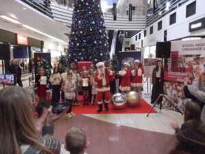 Grupa dzieci stoi obok Świętego Mikołaja i dwóch pomocnic na scenie w centrum handlowym, a w tle widoczna jest duża choinka.