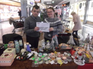 Para stoi za stołem, na którym prezentowane są rękodzieło i dekoracje. Trzymają kartkę z napisem: 'Stowarzyszenie "Źródło" - Warsztat Terapii Zajęciowej'.