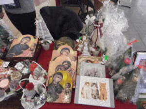 Stół eksponujący różne wyroby rękodzielnicze, w tym ikony religijne, małe figurki aniołów i stroiki choinkowe. Niektóre przedmioty są owinięte w celofan