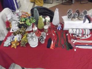 Na stole znajdują się różne ręcznie robione ozdoby świąteczne, w tym choinki z makaronów penne, ozdoby z szyszek sosnowych, dziergane akcesoria kwiatowe i kolorowe naszyjniki. Przedmioty są ułożone na czerwonym obrusie.