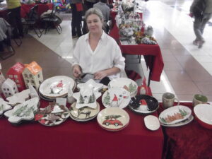 Kobieta siedzi za stołem, na którym eksponowane są świąteczne ceramiczne talerze w różnych kształtach: okrągłych, owalnych, w kształcie gwiazdek i choinek, przedstawiające choinki, skrzaty i bałwany, a także ceramiczne ozdoby na choinkę.