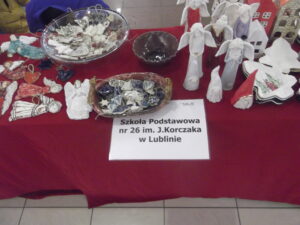 Stół nakryty czerwonym obrusem, na którym eksponowane są różne ręcznie robione wyroby ceramiczne i gliniane, takie jak ozdoby na choinkę oraz figurki aniołów i skrzatów. Na szyldzie widnieje napis Szkoła Podstawowa nr 26 im. J. Korczaka w Lublinie.