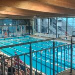 Kryty basen z wieloma torami, otoczony szklanymi ścianami i drewnianym sufitem. Widzowie siedzą po lewej stronie basenu