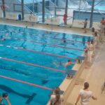 Jeden zawodnik trzyma się uchwytu basenu na mecie, a przed nim stoi lekko pochylona dziewczyna w białym stroju, trzymająca licznik czasu. Trzej pozostali zawodnicy płyną do mety, gdzie czekają na nich inne dziewczyny z licznikami.