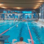 Kilku pływaków na torach podczas rozgrzewki. Po lewej stronie widzowie siedzą na trybunach.