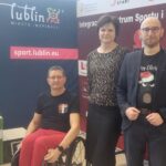 Kobieta i dwóch mężczyzn pozują przed banerami Integracyjnego Centrum Sportu i Rehabilitacji 'Start' oraz Lublina Miasta Inspiracji. Jedna osoba siedzi na wózku inwalidzkim obok podium. W tle znajduje się stół z wystawionymi trofeami sportowymi oraz upominkami w pudełku z logiem ICSiR 'Start' Lublin.