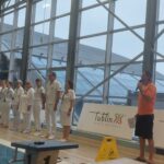 Grupa sędziów w białych strojach stoi w rzędzie w pobliżu basenu, zaś w niedalekiej odległości od nich stoi mężczyzna w pomarańczowej koszuli, który mówi do mikrofonu.