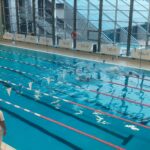 Trzech zawodników płynie w stronę mety, jeden daleko z tyłu za dwoma pozostałymi. Przy brzegu basenu stoi mężczyzna w białej koszulce z napisem 'sędzia' na plecach, obserwując przebieg zawodów