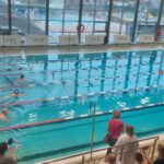 Zawodnicy pływacy na starcie, każdy z makaronikami pod pachami. Widok z trybun.