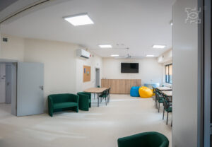 Jasny pokój z zielonymi kanapami, krzesłami przy stołach, pufami workami w kolorze żółtym i niebieskim, telewizorem na ścianie i tablicą ogłoszeniową.