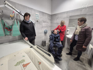 Cztery osoby w toalecie, jedna na wózku inwalidzkim. Jedna kobieta opiera się o podnośnik i leżankę, na której leży mini materac z kreskówkowym wzorem. Druga kobieta stoi za mężczyzną na wózku inwalidzkim. Trzecia kobieta spogląda na mężczyznę.