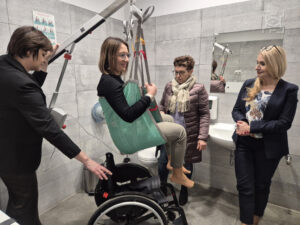 Cztery kobiety znajdują się w toalecie. Jedna kobieta siedzi na podnośniku nad wózkiem inwalidzkim, demonstrując jego użycie. Pozostałe kobiety stoją w pobliżu i obserwują.