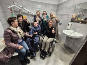 Siedem kobiet, w tym jedna na wózku inwalidzkim, oraz jeden mężczyzna na wózku inwalidzkim, pozują z uśmiechem do zdjęcia w środku toalety. Kilka osób podnosi kciuk w górę.