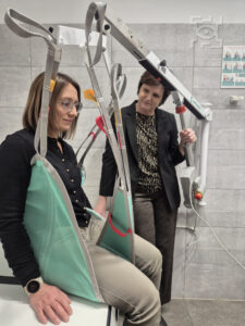 Dwie kobiety znajdują się w toalecie. Jedna siedzi w uprzęży podnośnika na leżance podczas gdy druga stoi w pobliżu i obserwuje. W tle widać kafelki i instrukcję obsługi na ścianie.