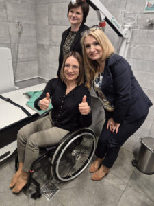 Trzy kobiety uśmiechają się przed zainstalowaną komfortką. Jedna kobieta siedzi na wózku inwalidzkim, pokazując kciuki do góry. Pozostałe dwie kobiety stoją obok niej, jedna pochyla się lekko do przodu. Znajdują się w toalecie z szarymi kafelkami na ścianach.