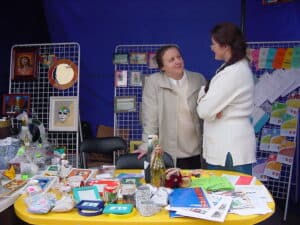 Dwie kobiety zwrócone w swoją stronę, stoją przy plastikowym żółtym stole, na którym ułożono rękodzieło i materiały informacyjne