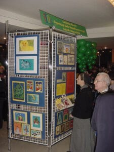 Kobieta w habicie oraz starszy mężczyzna oglądają prace konkursowe, wyeksponowane na metalowym stojaku