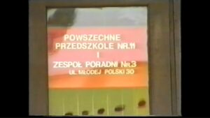 Kadr z filmu przygotowanego przez uczestników Pracowni Multimedialnej WTZ ze Świdnika; czerwona tabliczka z białym napisem "Powszechne Przedszkole nr 11 i Zespół Poradni nr 3 ul. Młodej Polski 30"