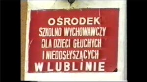 Kadr z filmu przygotowanego przez uczestników Pracowni Multimedialnej WTZ ze Świdnika; czerwona tabliczka z białym napisem "Ośrodek Szkolno-Wychowawczy dla Dzieci Głuchych i Niedosłyszących w Lublinie"