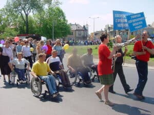 Na przedzie ulicznej parady miejskiej znajduje się troje pieszych z błękitnymi tablicami informacyjnymi. Napis na pierwszej tablicy brzmi: "Stowarzyszenie Osób Niepełnosprawnych Narządu Ruchu Promyk" napis na drugiej tablicy jest niekompletny. Następnie w kolejności znajduje się pięć osób jeżdżących na wózkach i ponownie duża grupa pieszych z balonami