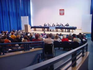 W centralnym punkcie auli przy długim stole siedzi sześć osób obu płci, z widowni obserwuje ich siedząca tyłem do fotografującego publiczność wśród której znajduje się mężczyzna na wózku