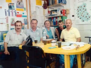 Zdjęcie grupowe: Kobieta, trzech mężczyzn i jeden młodzieniec siedzą wokół plastikowego żółtego stołu. Za nimi tablica korkowa z różnymi notatkami i emblematami oraz półka z segregatorami