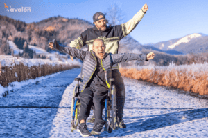 Mężczyzna w czapce z daszkiem z tyłu głowy i swetrze stoi za drugim mężczyzną na wózku inwalidzkim, na zaśnieżonej ścieżce. Obaj mają ręce uniesione w górę. W tle widać góry.