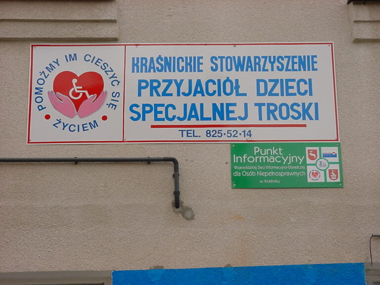Pod dużą białą tablicą z napisem "Kraśnickie Stowarzyszenie Przyjaciół Dzieci Specjalnej Troski, tel. 825 52 14"", umieszczono niewielką, zieloną tablicę z nazwą: "Punkt Informacyjny Wojewódzkiej Sieci Informacyjno-Doradczej dla Osób Niepełnosprawnych w Kraśniku