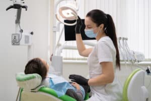 Dentystka w masce i rękawiczkach reguluje górne światło podczas badania pacjentki siedzącej na fotelu dentystycznym.