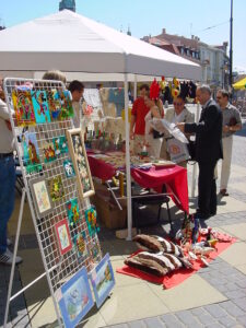 Przy stoisku zorganizowanym pod budynkiem Nowego Ratusza przy Placu Władysława Łokietka 1 w Lublinie w ramach projektu "Tęcza" stoją osoby oglądające prezentowane tu prace plastyczne: obrazy i rękodzieło