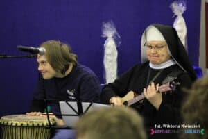 Od lewej siedzą: mężczyzna grający na bębenku i siostra zakonna grająca na gitarze