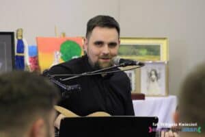 Przy mikrofonie siedzi grający na gitarze instruktor - Arkadiusz Sosnowski; w tle zanim wyeksponowano obrazy przeznaczone na aukcję